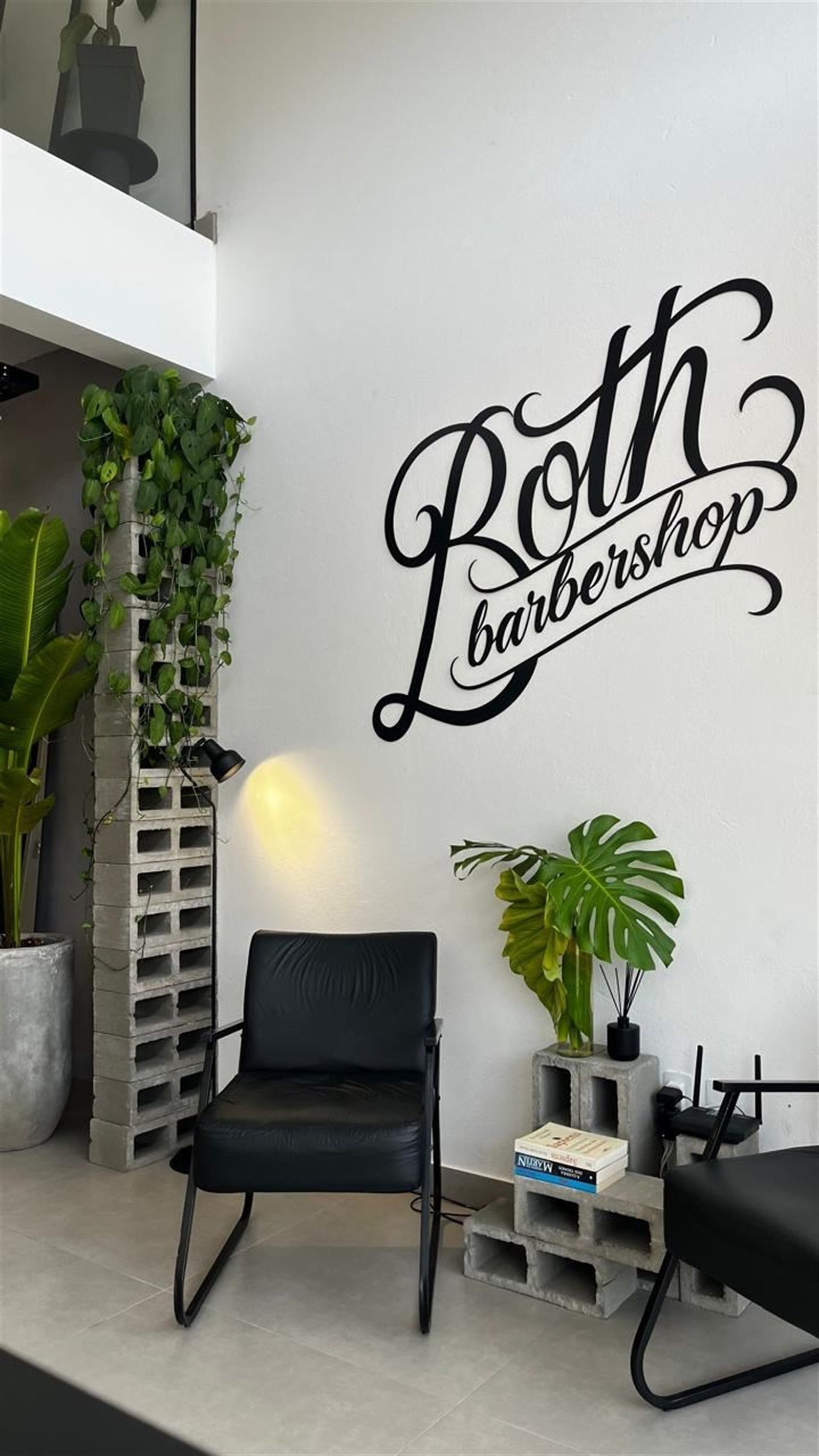 Parede assinada Both Barbershop, com poltronas de espera em couro preto e pilar de blocos de cimento com plantas
