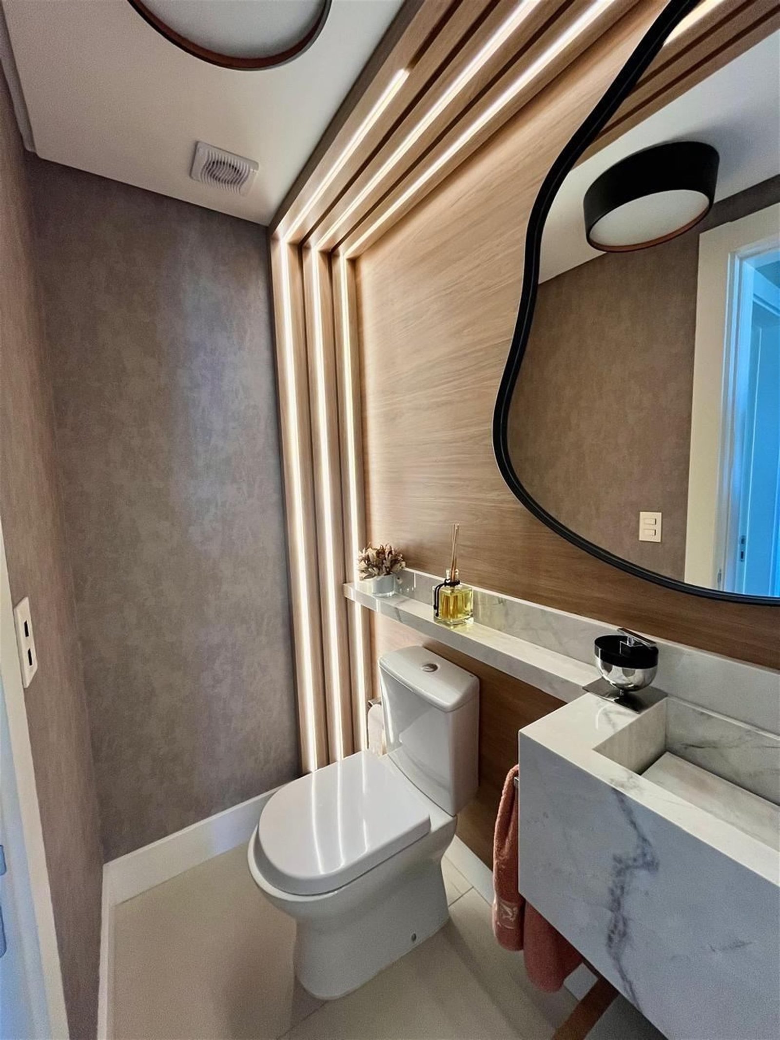 Lavabo com painel amadeirado ripado, espelho de formato orgânico e bancada em pedra clara